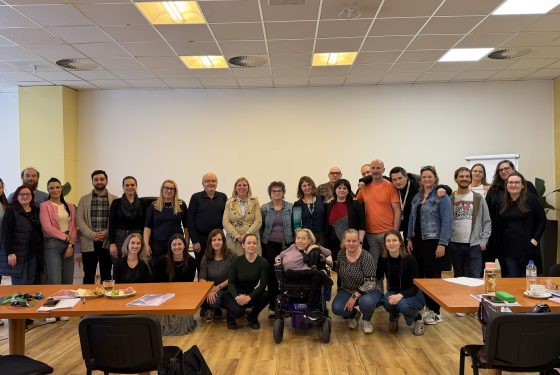 Skupinska fotografija udeleženk in udeležencev usposabljanja NDTi 2026 v seminarski dvorani. Skupina stoji in sedi za mizami v svetlem prostoru.